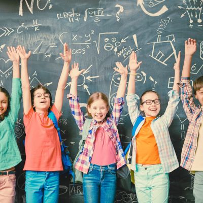 Children with their hands up standing in front of a chalkboard experiencing school success