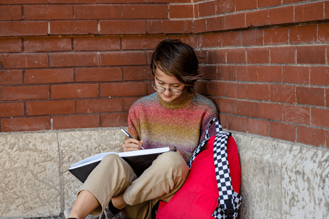 young girl journalling outside leaning against a brick wall.